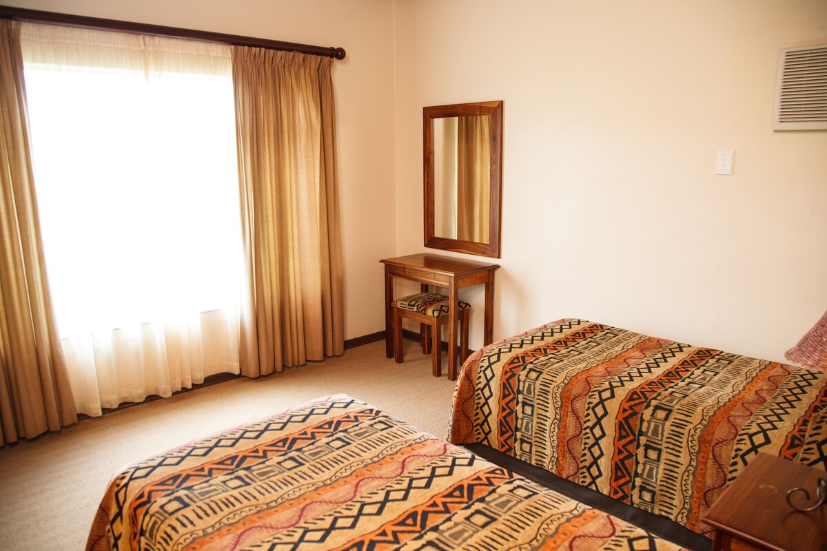 A room with two beds, a wooden chair, a mirror, and a window with curtains.