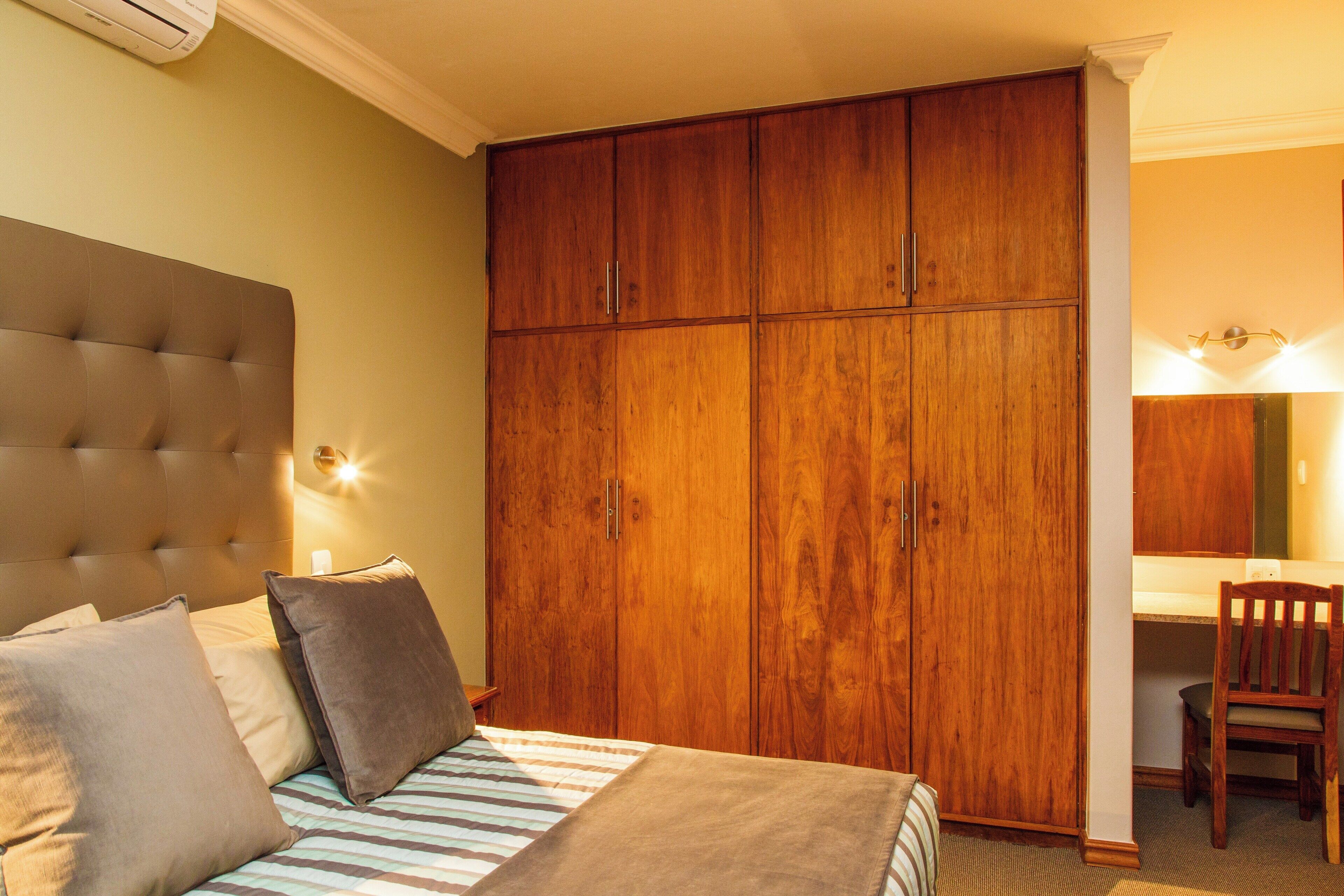 Une chambre à coucher avec une grande armoire en bois, une tête de lit capitonnée et une table de chevet avec une lampe.
