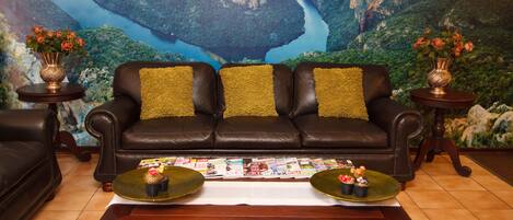 A living room with a large mural of a canyon, a leather sofa with green cushions, and two side tables with vases of flowers.