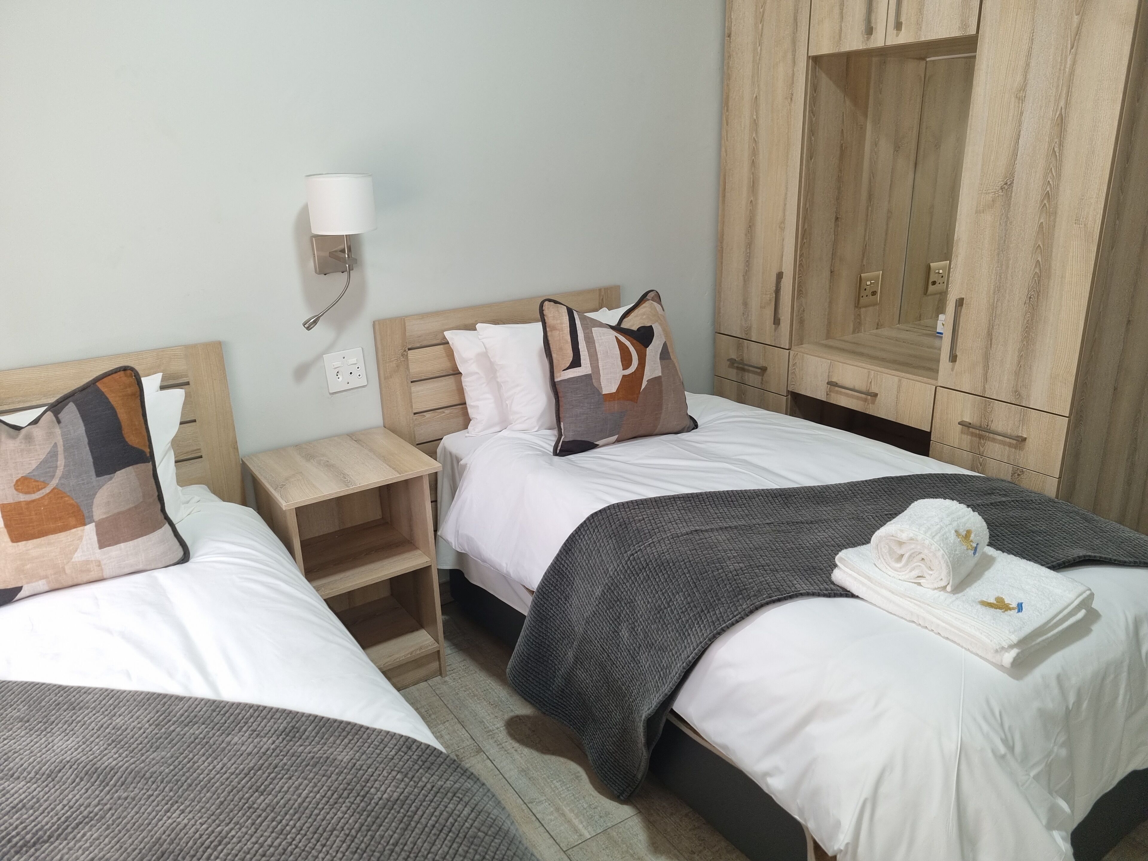 A hotel room with two beds, wooden headboards, a nightstand, a wardrobe, and a wall-mounted lamp.