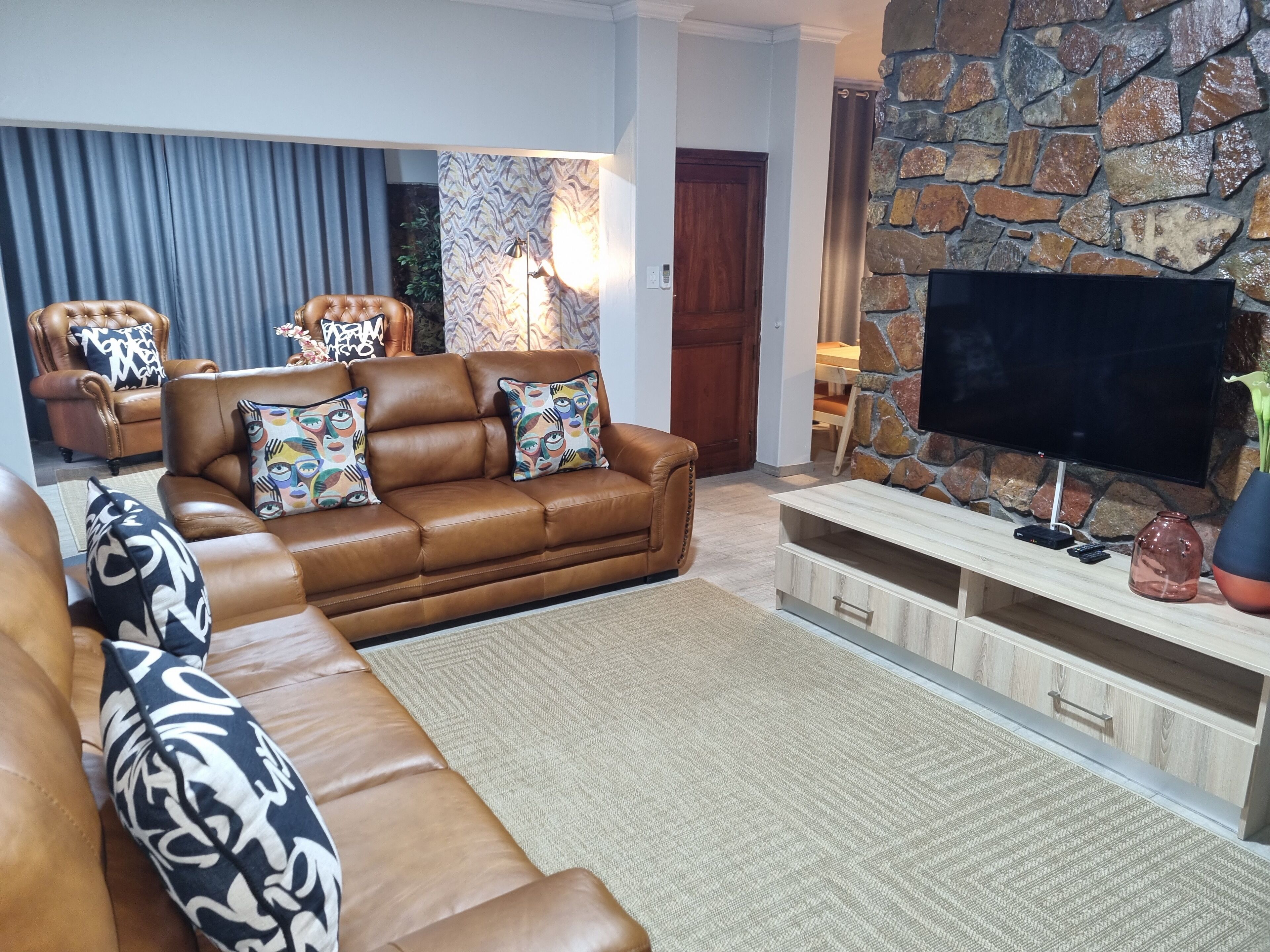 A living room with a brown leather sofa, patterned cushions, a flat-screen TV, and a stone wall.