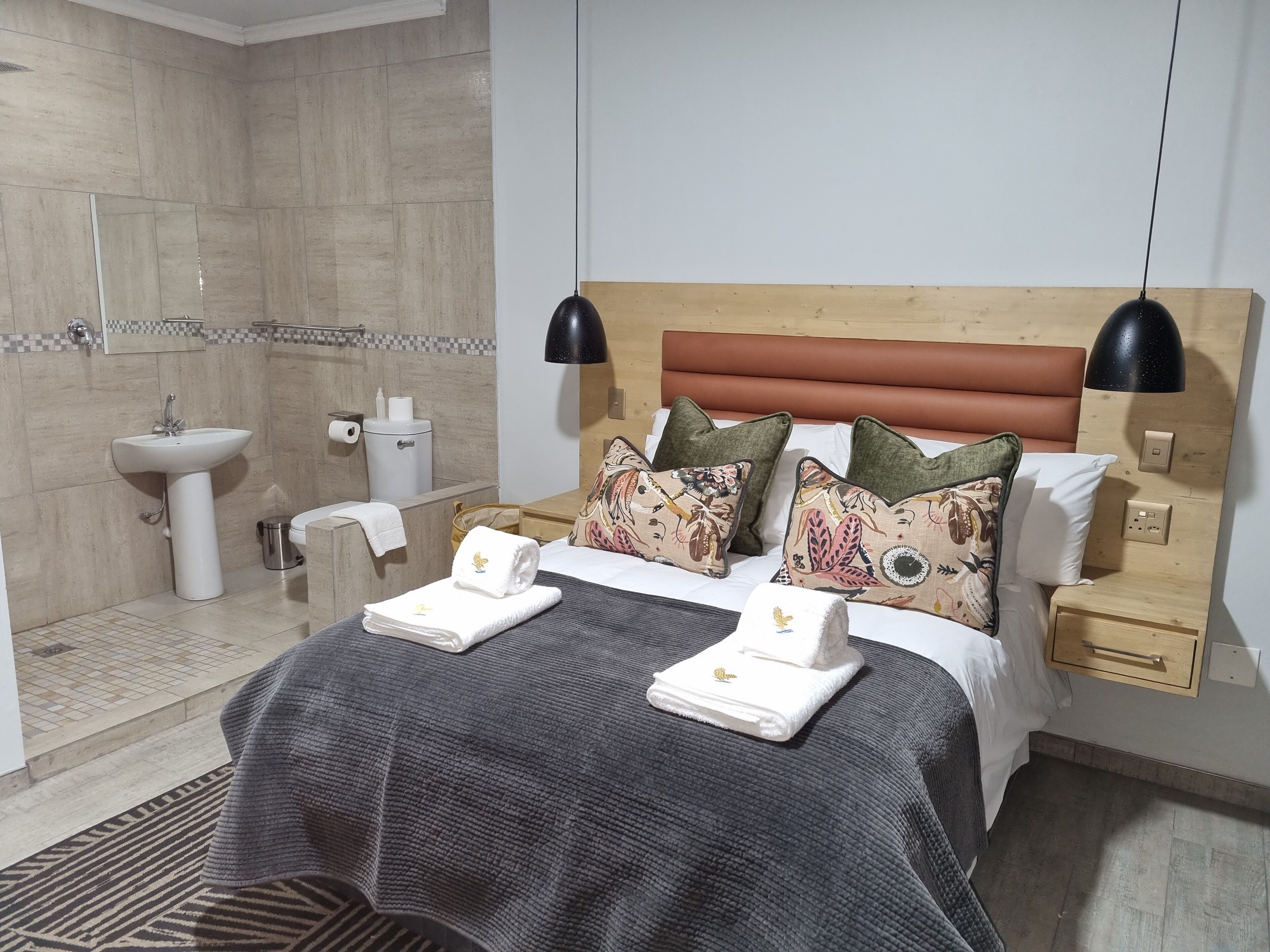 A hotel room with a bed, two bedside tables, a nightstand, a wooden headboard, a wall-mounted lamp, a bathroom with a sink and toilet, and a patterned rug.