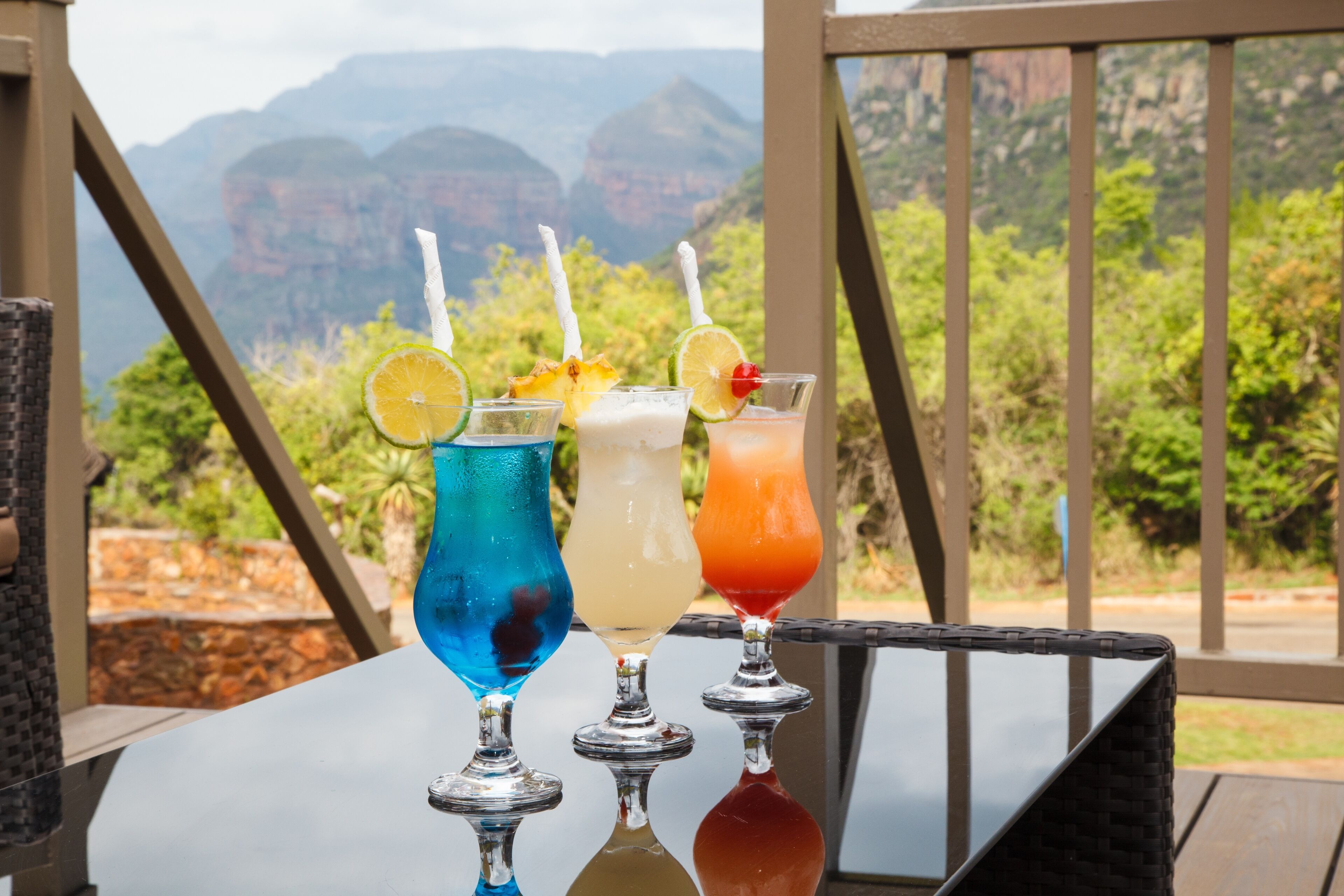 Tres cócteles coloridos sobre una mesa, con una vista panorámica de montañas al fondo.
