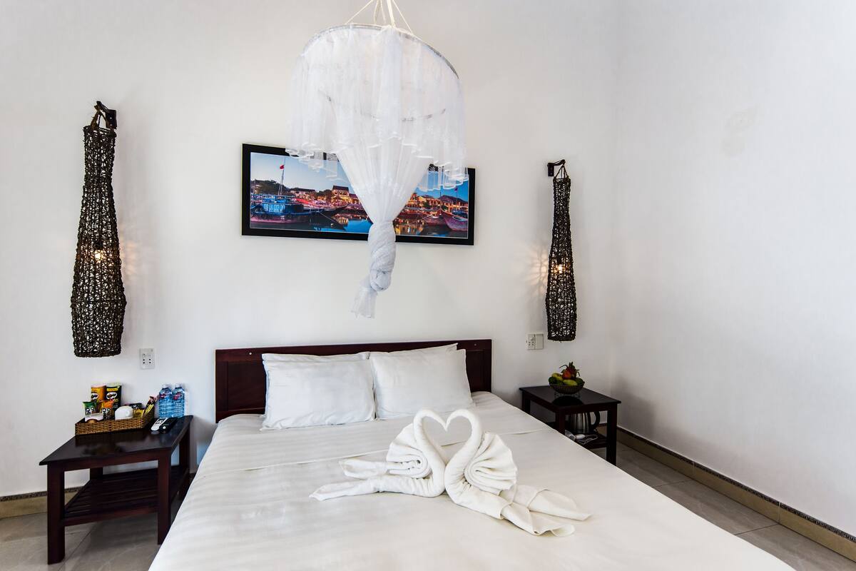 A hotel room with a neatly made bed, a framed picture on the wall, and a small table with a basket and a fruit bowl.