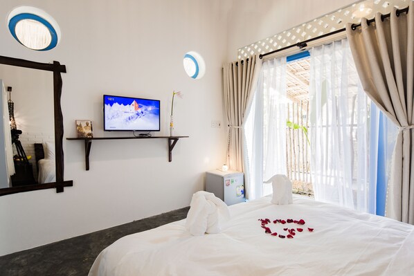 A hotel room with a bed, a TV, a small desk, a mirror, and a window with curtains. - Cashew Tree Bungalow (Da Nang)