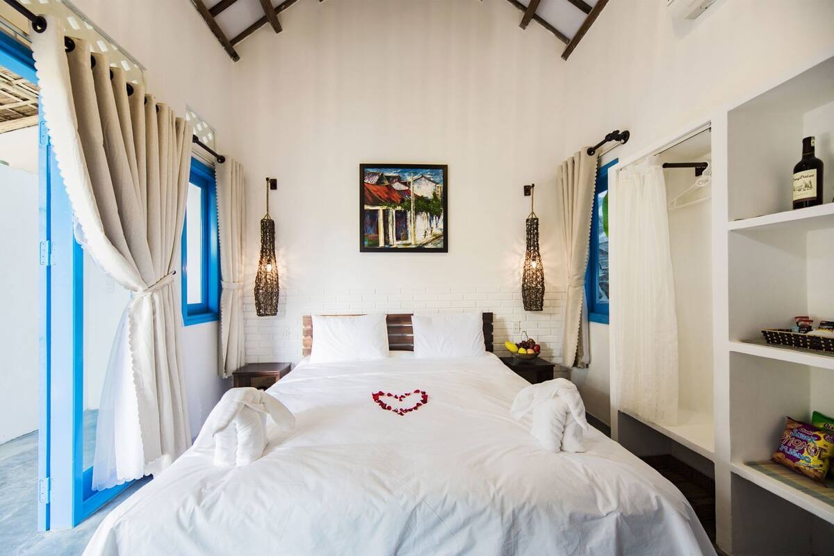 A hotel room with a bed, a painting, a bottle of wine, and a fruit bowl.