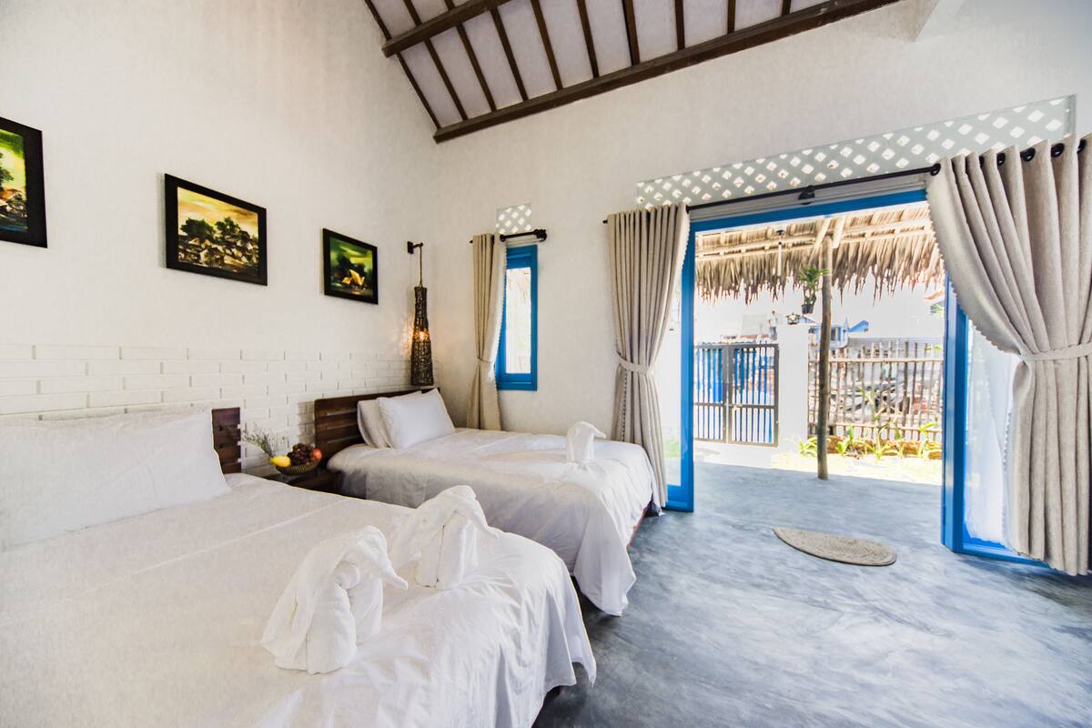 A hotel room with two beds, a balcony with a thatched roof, and a view of the outside.
