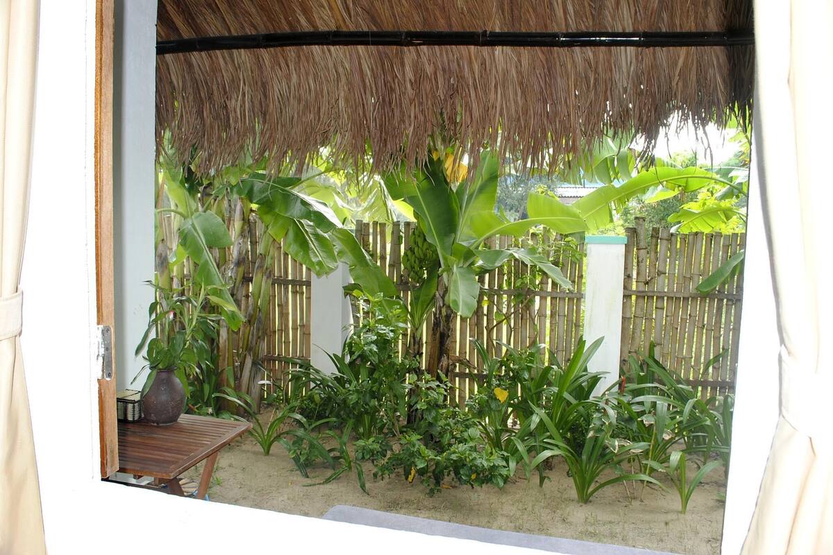 A thatched-roof structure with a wooden table and a potted plant, surrounded by lush greenery and a bamboo fence.