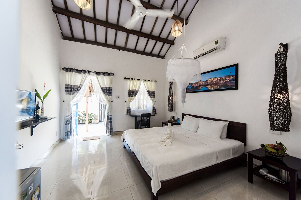 A bedroom with a bed, bedside tables, a desk, a chair, a hanging light fixture, a wall-mounted air conditioner, and a painting.