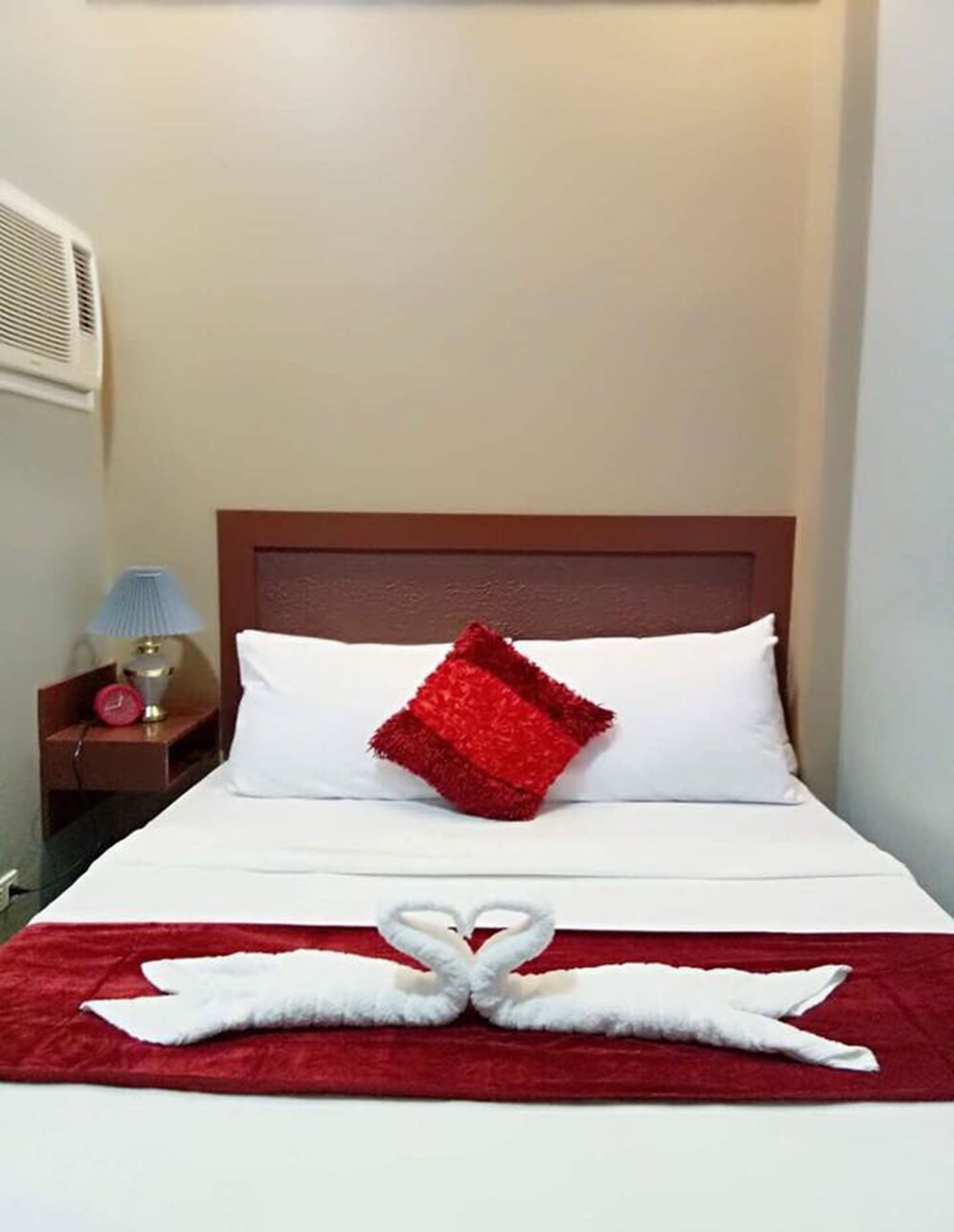 A hotel room with a bed, a red pillow, a red blanket, a heart-shaped towel arrangement, a nightstand with a lamp, and a wall-mounted air conditioner.