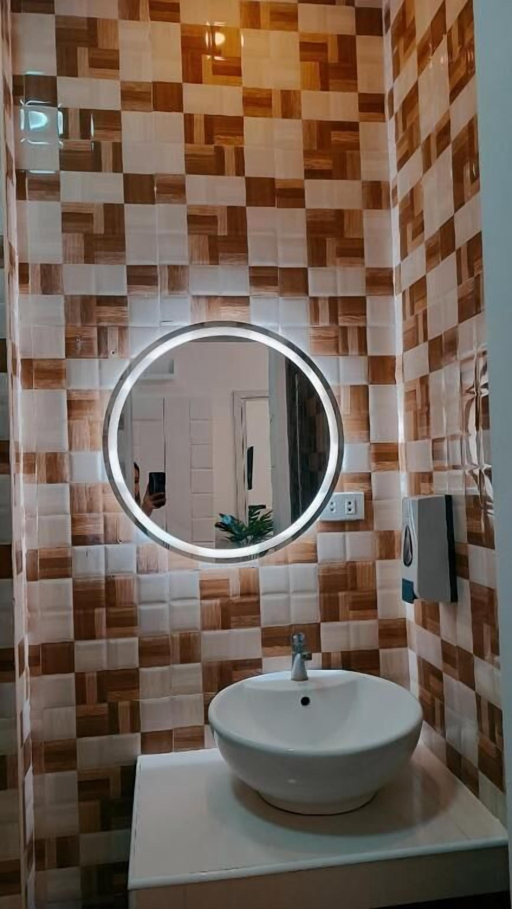 Um banheiro com painel de azulejos xadrez, um espelho redondo com iluminação e uma pia branca.
