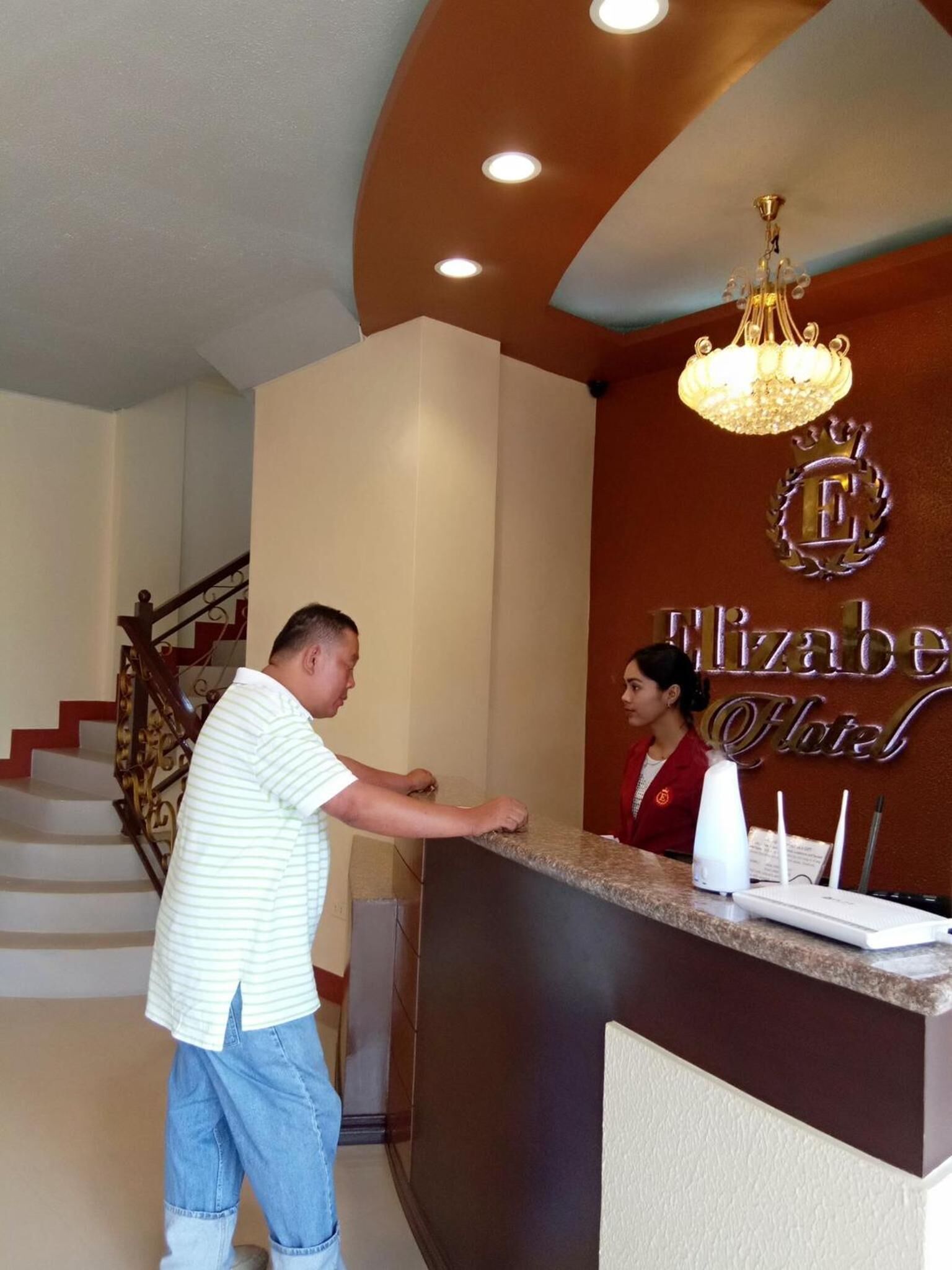 A hotel reception area with a receptionist, a reception desk, a chandelier, and a staircase in the background.