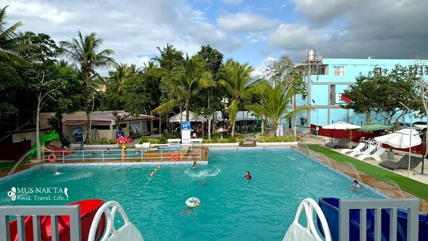 En swimmingpool med rutsjebane, omgivet af palmer og en bygning med et skilt, der siger "MUS NAK'TA Food. Travel. Life.".