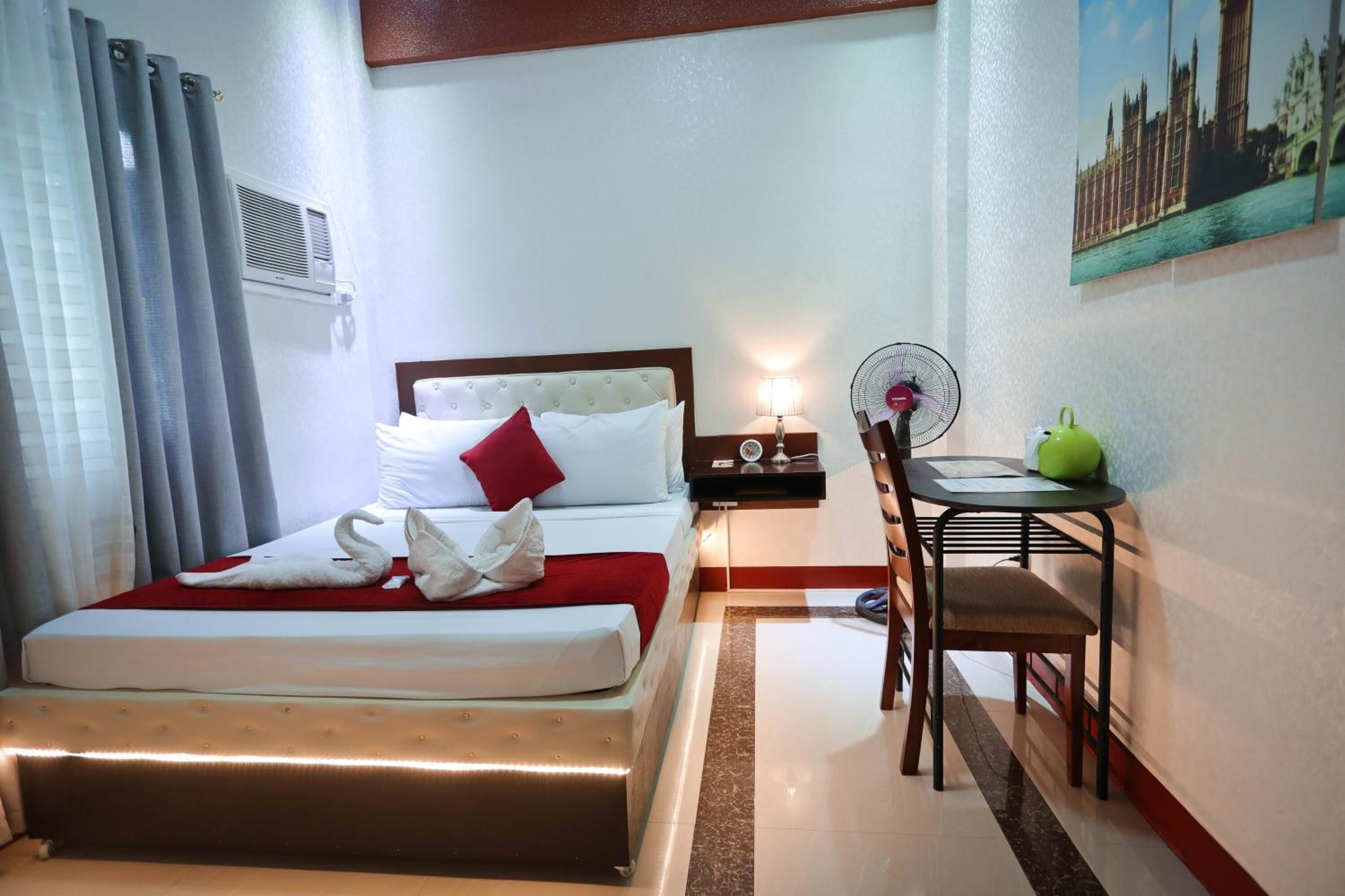 Una habitación de hotel con cama, escritorio, silla, ventilador y un cuadro en la pared.