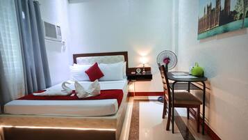 Una habitación de hotel con cama, escritorio, silla, ventilador y un cuadro en la pared.