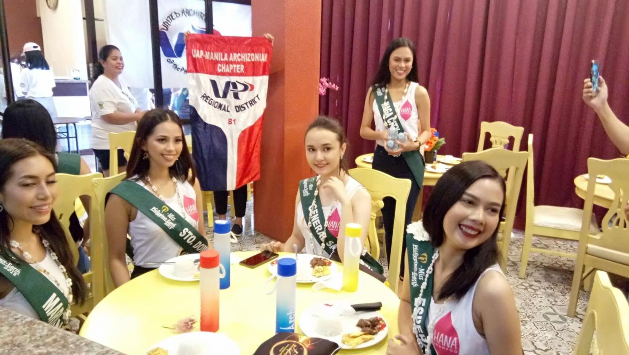 A group of people seated at a round table with plates of food, in a room with a red curtain backdrop and a banner for the UAP-Manila Archi-