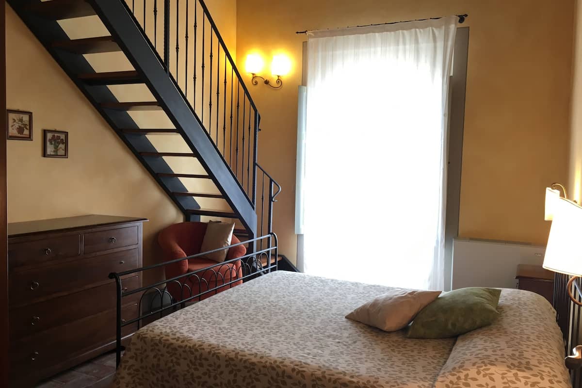 Une chambre à coucher avec un lit, une commode en bois, une chaise, un escalier et une fenêtre avec des rideaux.