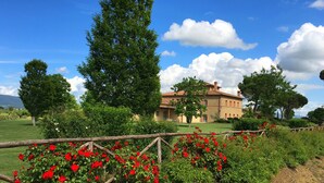 Un grand bâtiment en pierre, surmonté d’un toit de tuiles, entouré d’une végétation luxuriante et de fleurs rouges.