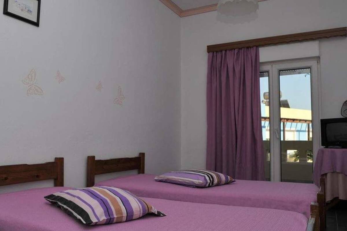 A room with two beds, a window with purple curtains, a fan, and a small table with a tablecloth.