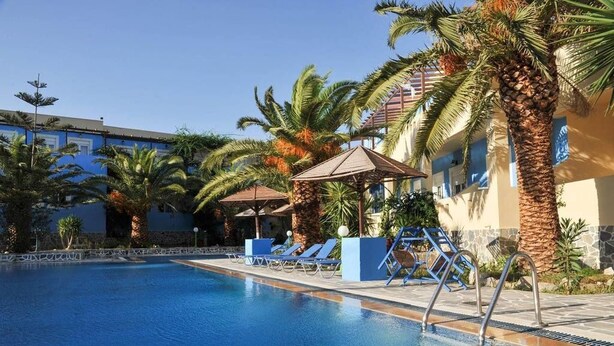 Una piscina con sillas de descanso y sombrillas, rodeada de palmeras y un edificio de paredes azules y beige.