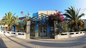 Un edificio con entrada protegida por una reja, rodeado de palmeras y plantas con flores.