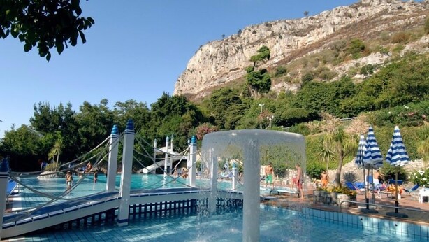 A swimming pool with a water fountain, surrounded by umbrellas and a rocky hillside in the background. - Holiday Time Sorrento (Piano di Sorrento)