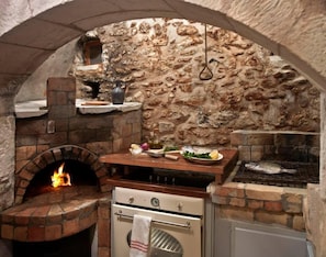 An outdoor kitchen with a stone wall, a brick oven, a grill, and a wooden counter with various cooking utensils and ingredients.