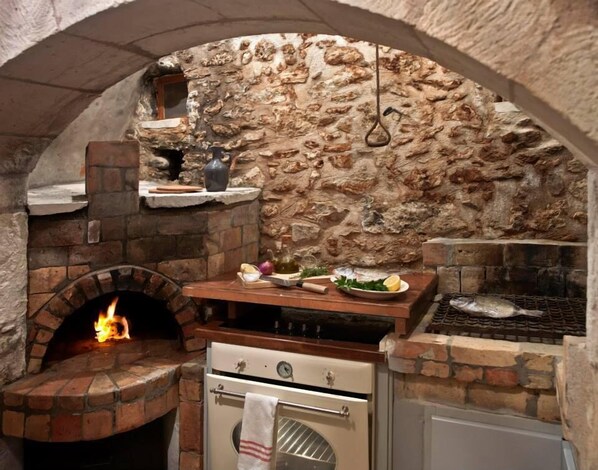 An outdoor kitchen with a stone wall, a brick oven, a grill, and a wooden counter with various cooking utensils and ingredients.