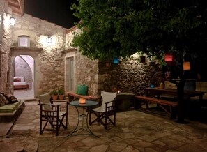 An outdoor patio area with stone walls, a wooden bench, a table with chairs, and a bed visible through an archway. - Critamo Cottage (Apokoronas)