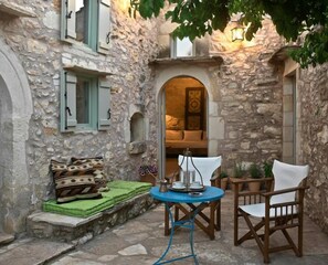 A stone-walled courtyard with a round table and two chairs, a cushioned bench, and a small archway leading to an interior room.