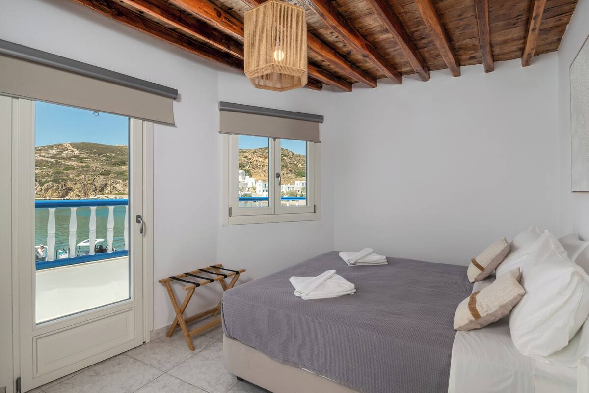 Un dormitorio con cama, ventana con vista al mar, balcón con una silla y techo de madera.