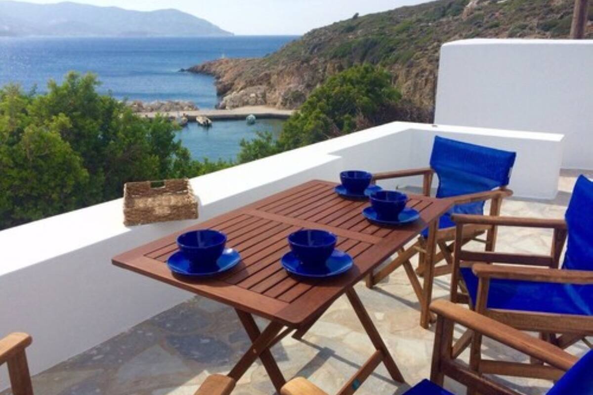 Una mesa de madera con sillas azules y cuencos en una terraza con vistas al mar y a las montañas.