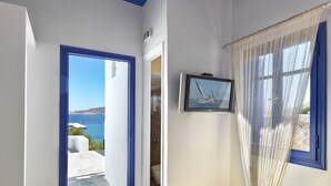 A room with a bed, a television on the wall, a view of the sea, and a bathroom door. - Psathi Blue (Kimolos)