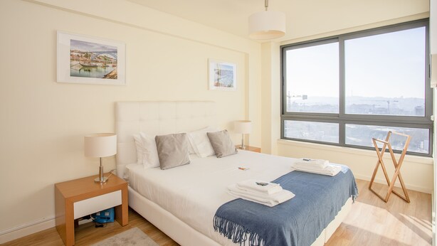 Une chambre d’hôtel moderne avec un grand lit, une table de chevet avec une lampe, une fenêtre donnant sur la ville et deux tableaux encadrés au mur.