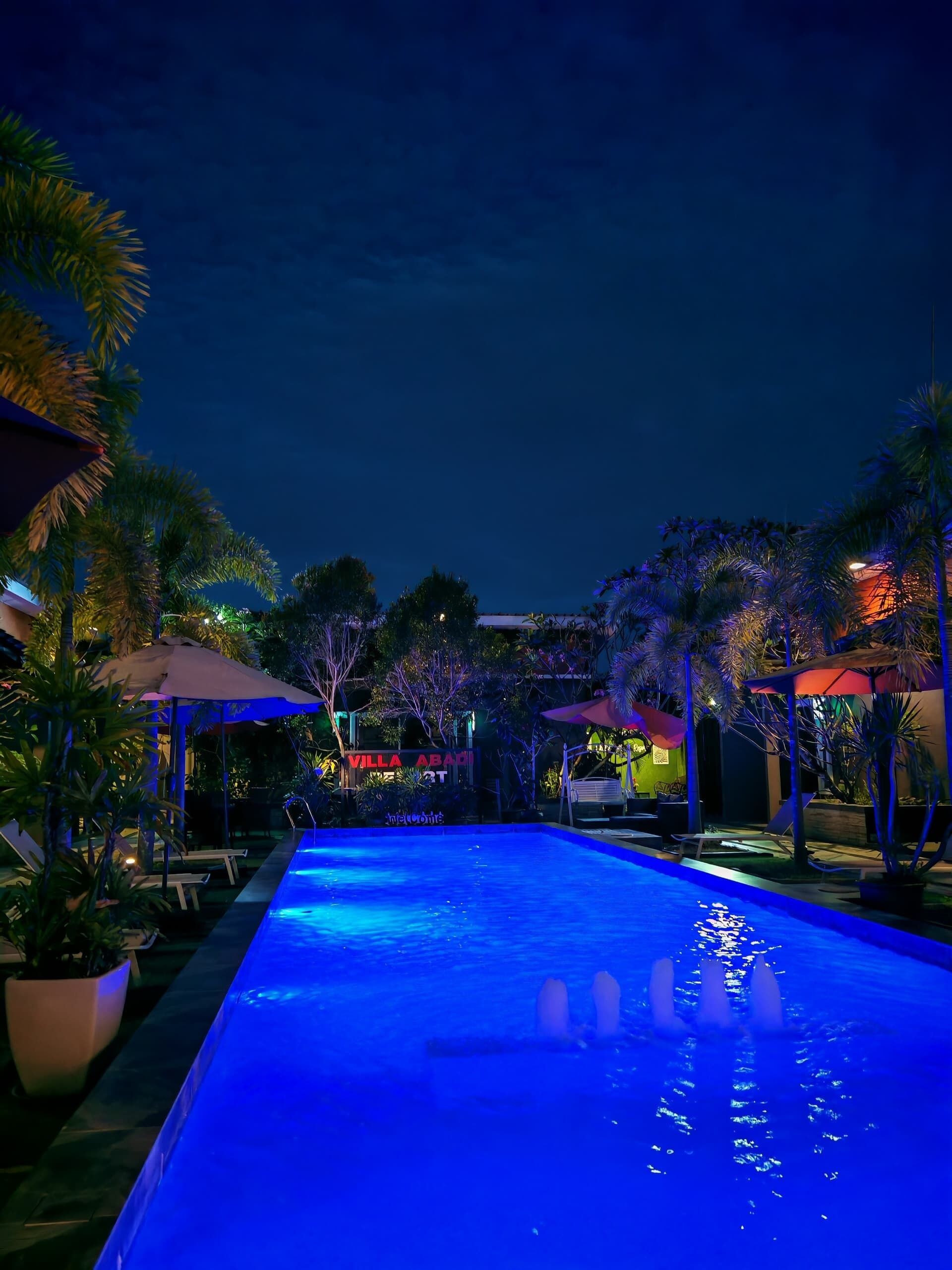 Una piscina con sillas de descanso y sombrillas, rodeada de palmeras y un edificio con señalización iluminada.