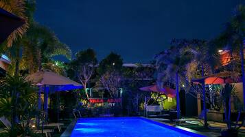 Una piscina con sillas de descanso y sombrillas, rodeada de palmeras y un edificio con señalización iluminada.