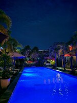 Una piscina con sillas de descanso y sombrillas, rodeada de palmeras y un edificio con señalización iluminada.
