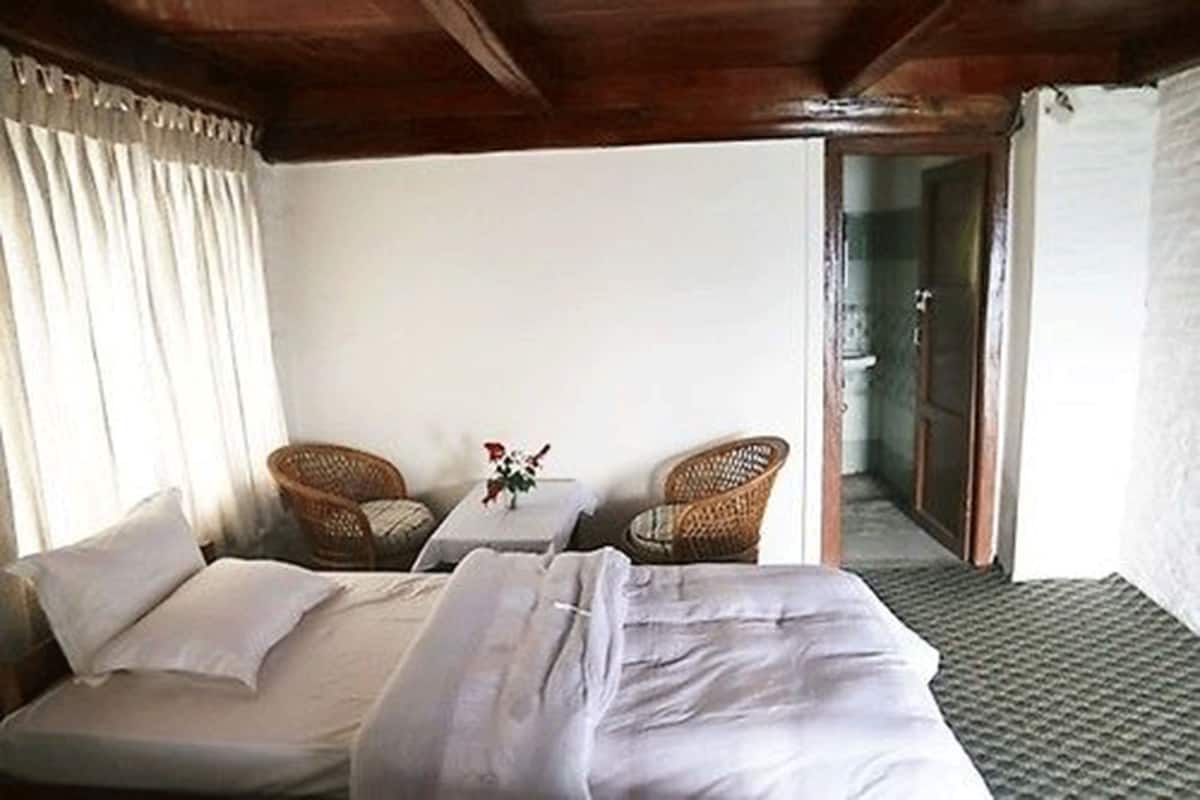 A room with a bed, two wicker chairs, a small table with a vase, and a view of a bathroom.