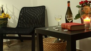 A room with a black chair, a table with a book, a bottle, two glasses of wine, a candle, and a basket of magazines.