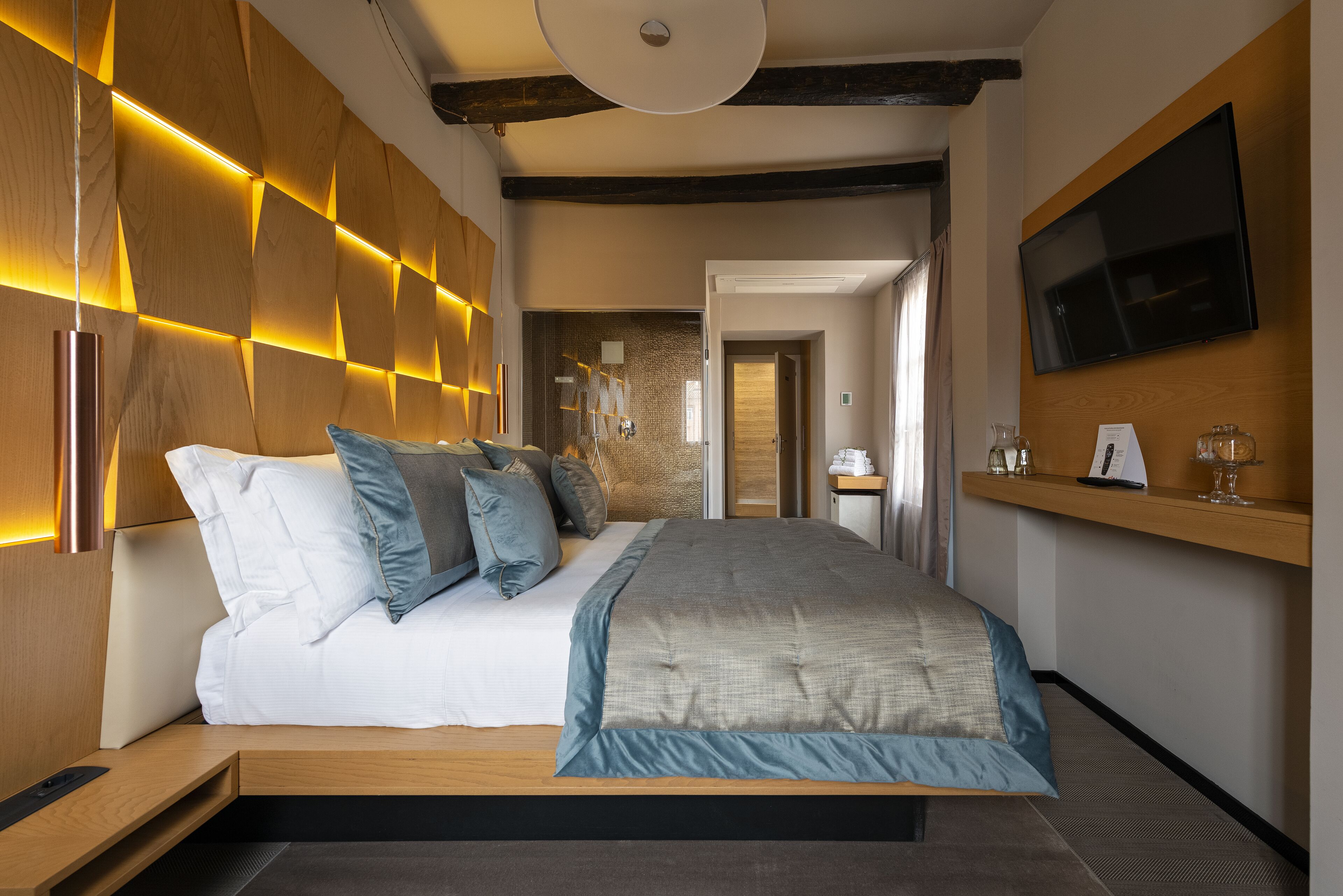 A modern hotel room with a large bed, wooden headboard, bedside tables, a flat-screen TV, and a small shelf with glasses.