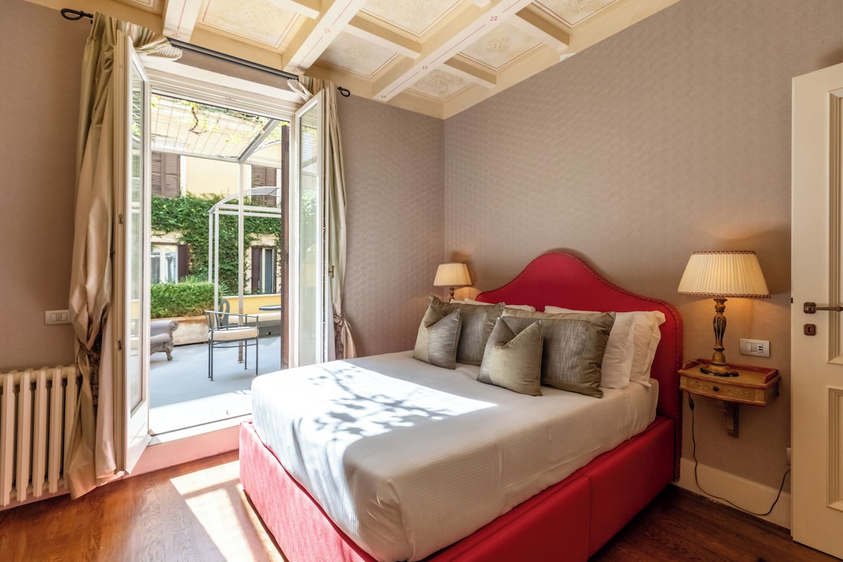 A bedroom with a red bed, two bedside lamps, a view through an open door to an outdoor area with a table and chairs.