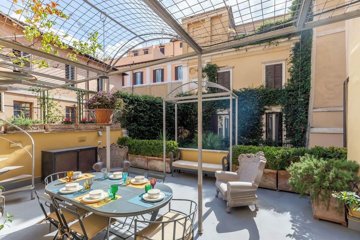 Patio esterno luminoso con tavolo rotondo apparecchiato per mangiare, circondato da comodi posti a sedere e piante in vaso.