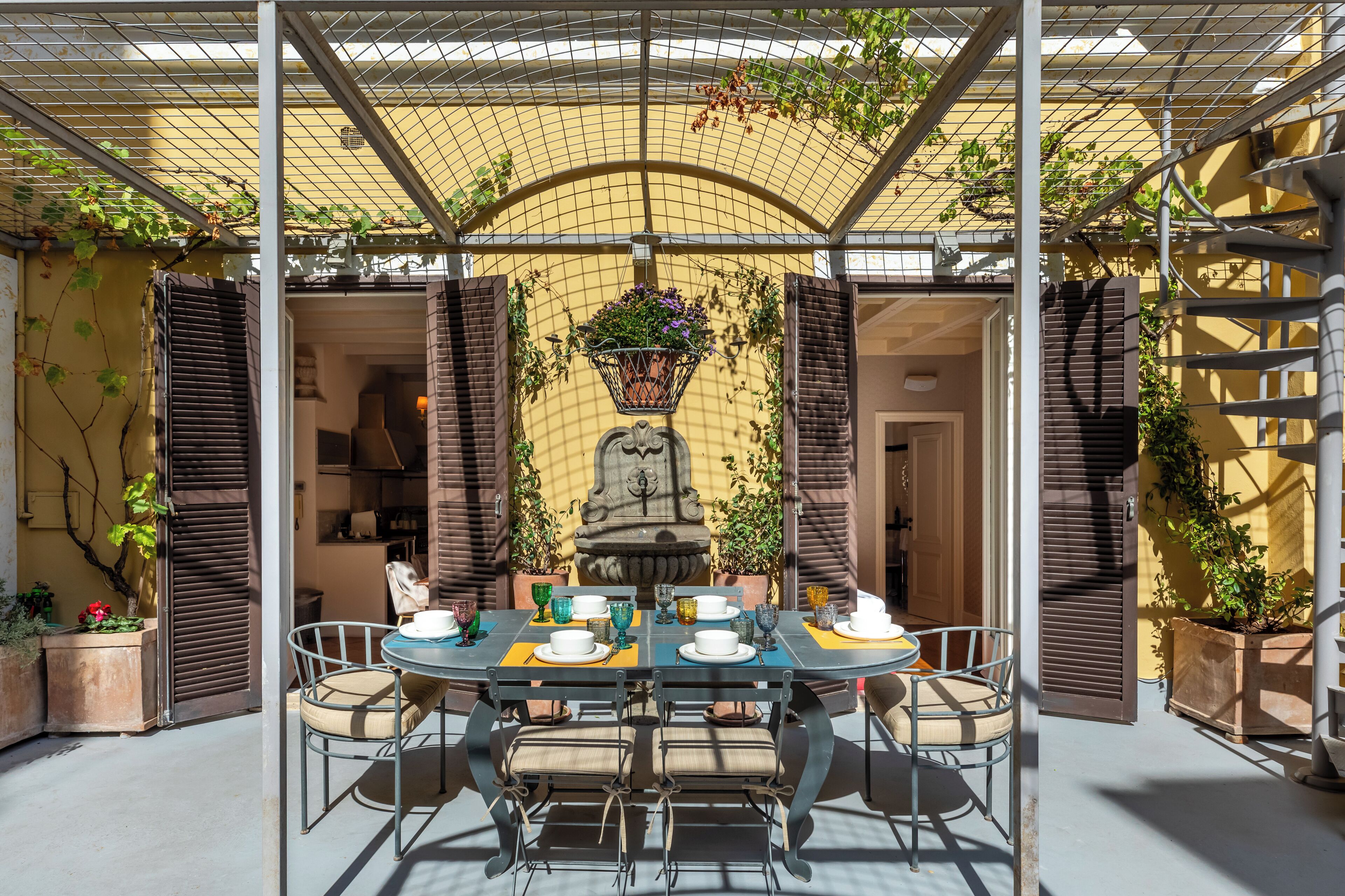 Un área de comedor al aire libre cubierta con una mesa dispuesta para seis personas, rodeada de sillas y una estatua al fondo.