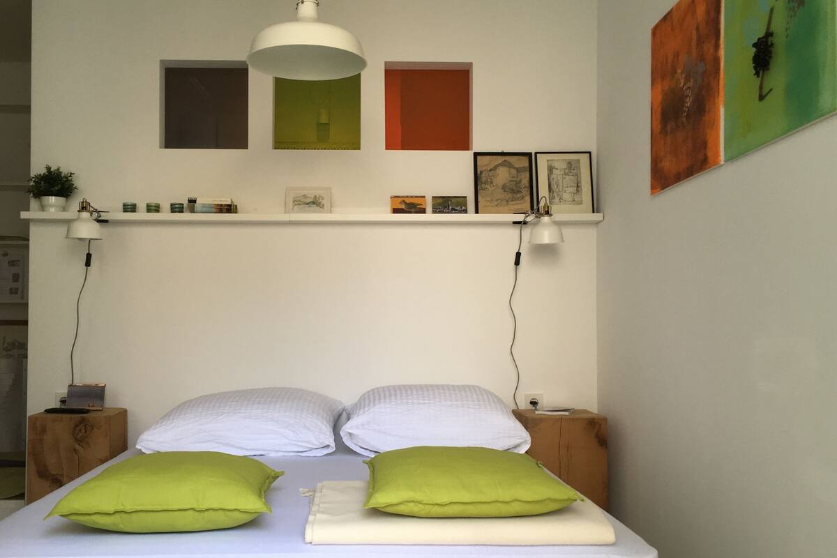Una cama con dos almohadas, una mesita de noche, una estantería con libros e imágenes, y una lámpara colgante.