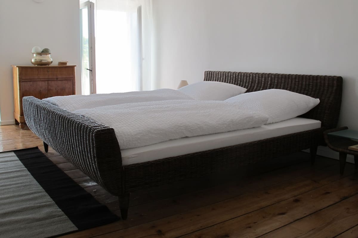 Una cama doble con colchón y almohadas blancas, un cabecero de mimbre, un aparador de madera y una mesita de noche con lámpara.