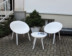 Two white chairs and a small table with a magazine on it, placed on a paved area with a potted plant nearby. - Quartier31 (Lieser)