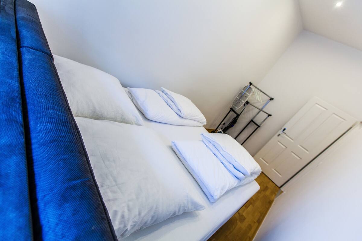 A bedroom with a bed, pillows, a folded towel, a wall-mounted heater, and a door.