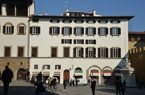 Une place publique avec un grand bâtiment doté de nombreuses fenêtres et portes, une statue et quelques personnes qui se promènent.
