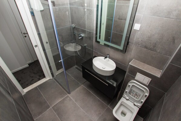 A modern bathroom with a glass-enclosed shower, a white sink, and a toilet with a digital display. - Hotel Alkima (Kotor)