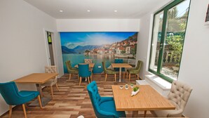 A dining area with wooden tables and chairs, a mural of a coastal town, and a large window with a view of greenery. - Hotel Alkima (Kotor)